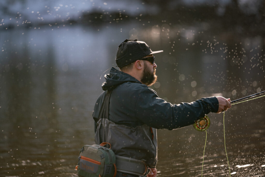 A young man fly fishing during the winter in the Upper Peninsula wearing a black hat, blue jacket, and gray waders.